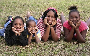Photograph of four African American siblings laying on their stomachs on a lawn side by side with their heads propped up on hands and elbows smiling